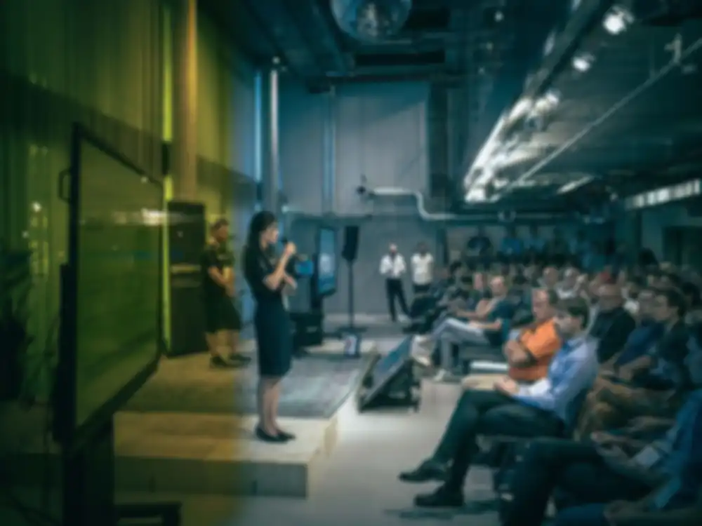 A woman stands on stage at a conference and speaks to interested participants