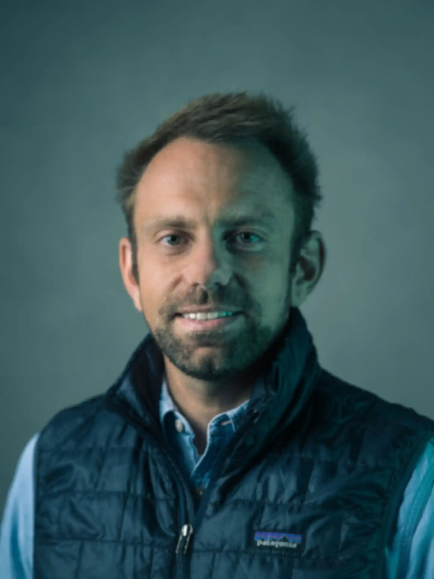 A portrait photo of Christopher Kränzler, standing in front of a plain wall and smiling at the camera. He is wearing a down vest, and the photo is bathed in a greenish light.