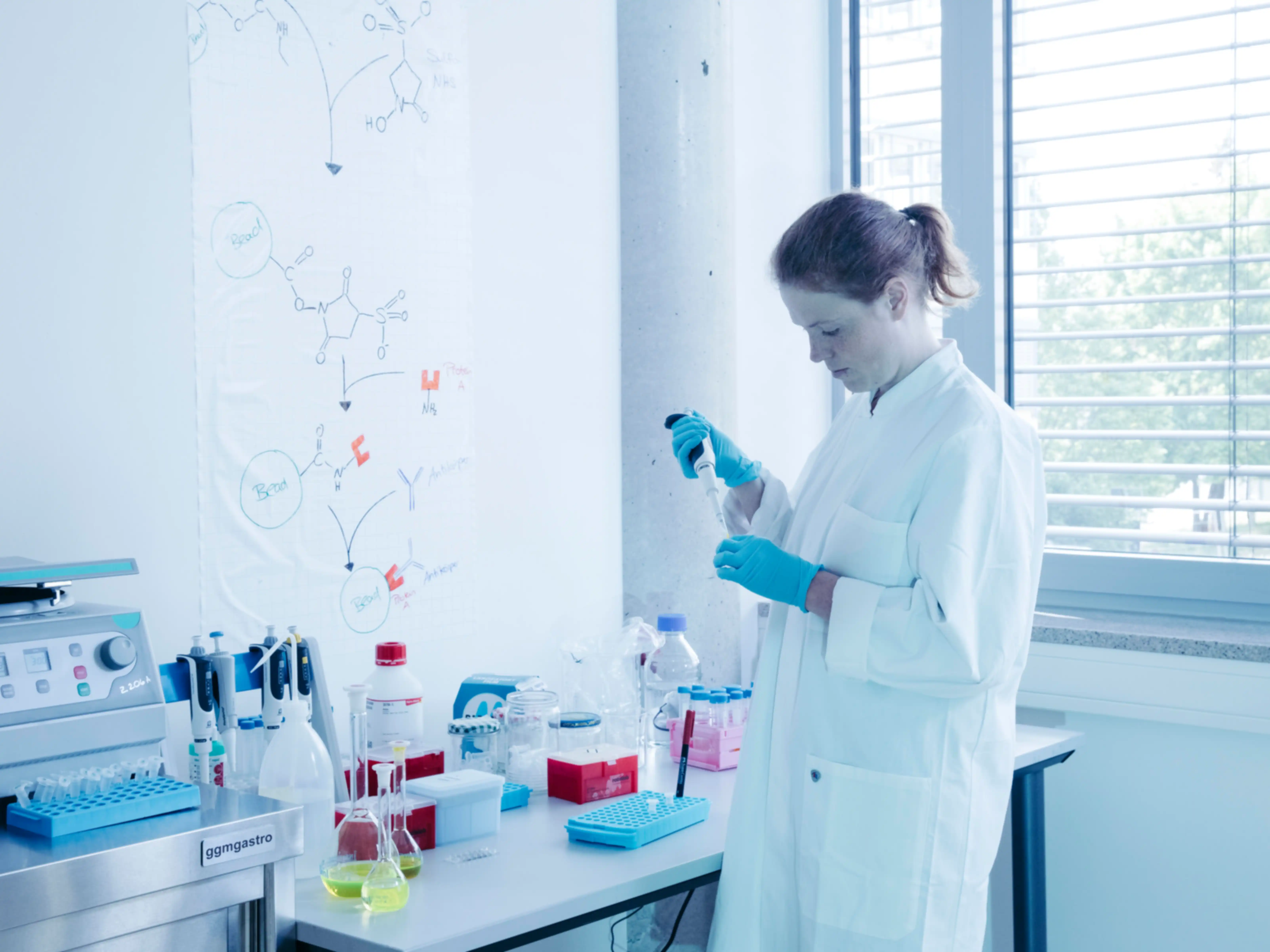 A woman wearing a lab coat fills a container with a pipette in a laboratory setting
