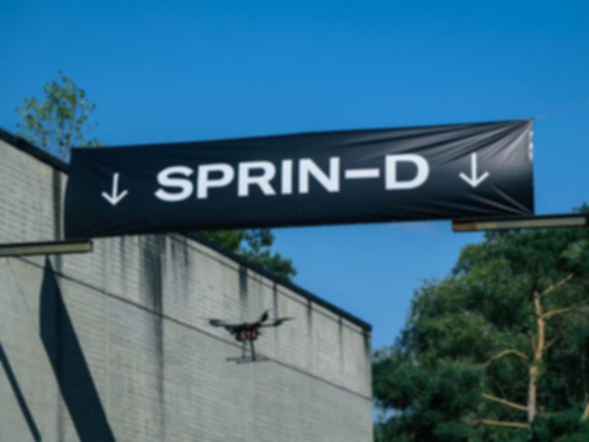 Eine Drone fliegt autonom zwicschen zwei Hangars und unter einem Banner hindurch mit der Aufschrift SPRIND